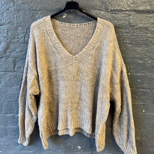 May include: A beige, oversized V-neck sweater hangs on a black hanger against a gray brick wall. The sweater has a textured knit design across the chest and a ribbed cuff and hem. The sleeves are long and the fit is loose.