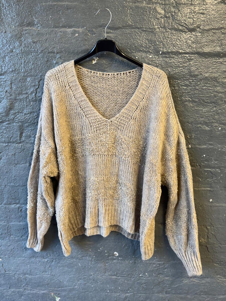 May include: A beige, oversized V-neck sweater hangs on a black hanger against a gray brick wall. The sweater has a textured knit design across the chest and a ribbed cuff and hem. The sleeves are long and the fit is loose.