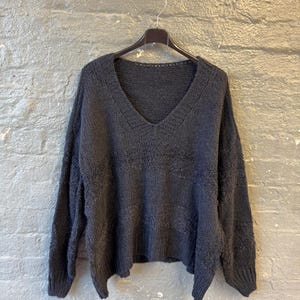 May include: Dark gray, oversized sweater with a deep V-neck. The sweater is made of a soft, textured knit fabric and hangs on a black hanger against a gray brick wall. The sleeves are long and the fit is loose.