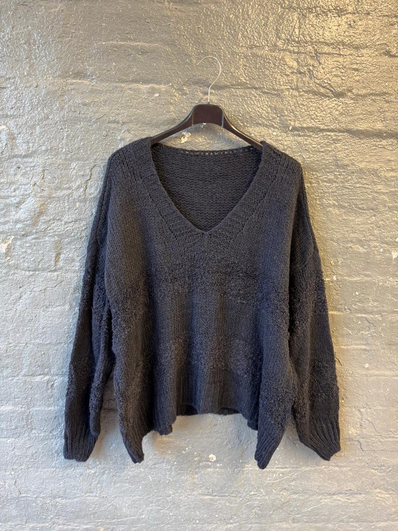 May include: Dark gray, oversized sweater with a deep V-neck. The sweater is made of a soft, textured knit fabric and hangs on a black hanger against a gray brick wall. The sleeves are long and the fit is loose.