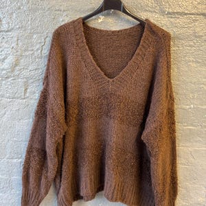 May include: A brown, V-neck sweater hangs on a black hanger against a gray brick wall. The sweater has a textured knit design and long sleeves. The color is a warm, earthy brown.