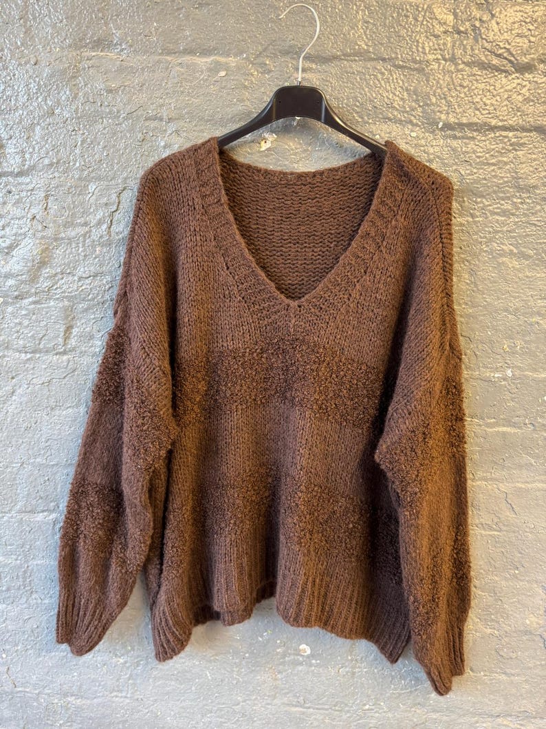 May include: A brown, V-neck sweater hangs on a black hanger against a gray brick wall. The sweater has a textured knit design and long sleeves. The color is a warm, earthy brown.