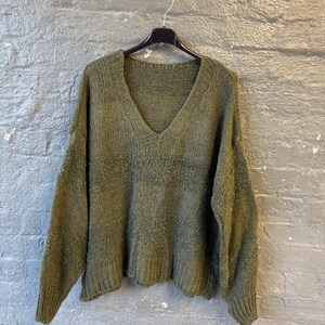 May include: Olive green knit sweater with a V-neck. The sweater is hanging on a black hanger against a gray brick wall. The knit has a textured appearance.