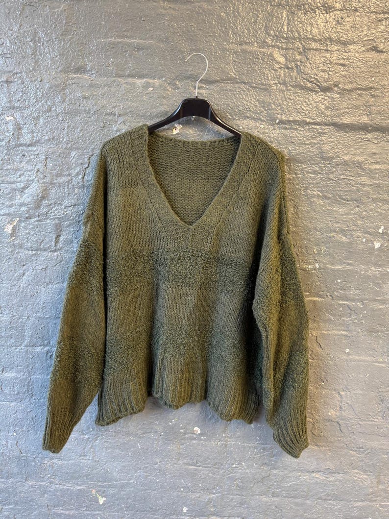 May include: Olive green knit sweater with a V-neck. The sweater is hanging on a black hanger against a gray brick wall. The knit has a textured appearance.