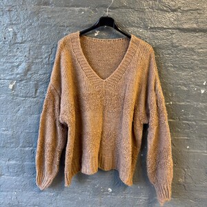 May include: A tan-colored, V-neck sweater hangs on a black hanger against a gray brick wall. The sweater has long sleeves and a textured knit pattern. The neckline and cuffs are ribbed.