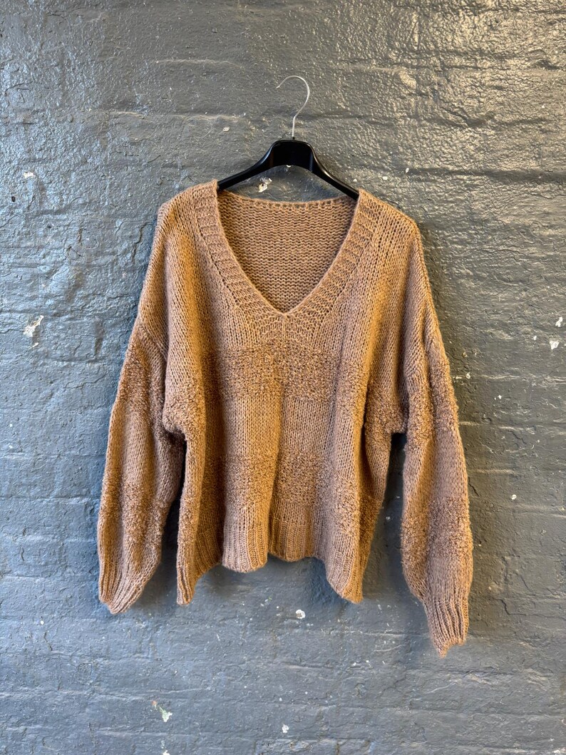 May include: A tan-colored, V-neck sweater hangs on a black hanger against a gray brick wall. The sweater has long sleeves and a textured knit pattern. The neckline and cuffs are ribbed.