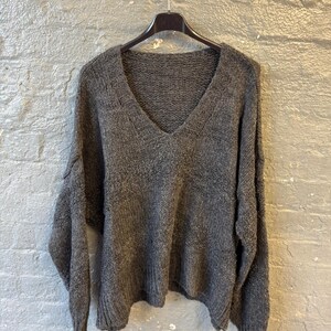 May include: A dark gray, V-neck sweater hangs on a black hanger against a textured gray brick wall. The sweater has long sleeves and a loose, comfortable fit. The knit fabric appears soft and slightly textured, perfect for cooler weather.