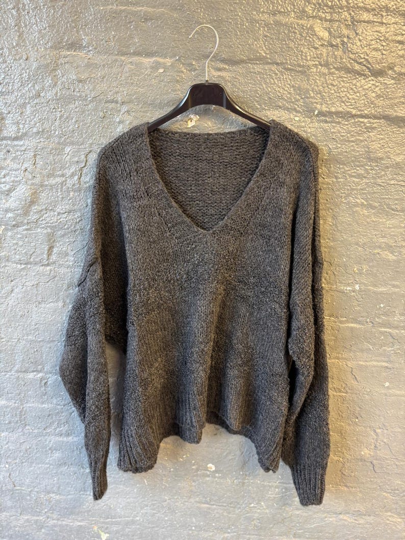 May include: A dark gray, V-neck sweater hangs on a black hanger against a textured gray brick wall. The sweater has long sleeves and a loose, comfortable fit. The knit fabric appears soft and slightly textured, perfect for cooler weather.