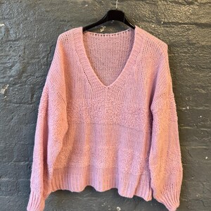 May include: A light pink, V-neck sweater with long sleeves hangs on a black hanger. The sweater has a textured knit design across the chest and a ribbed hem and cuffs. The background is a dark gray brick wall.