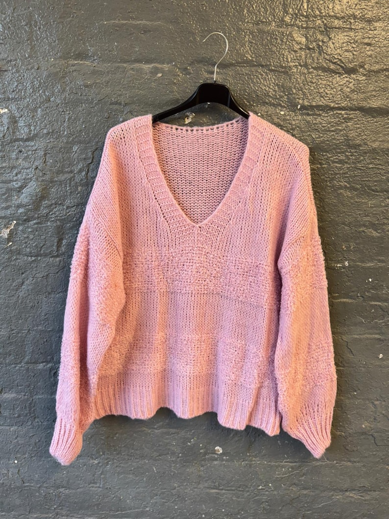 May include: A light pink, V-neck sweater with long sleeves hangs on a black hanger. The sweater has a textured knit design across the chest and a ribbed hem and cuffs. The background is a dark gray brick wall.