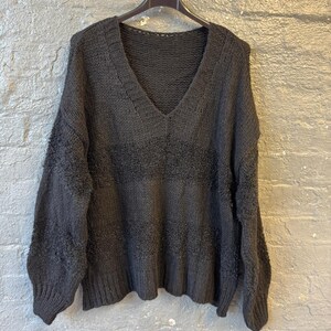 May include: A dark gray, oversized sweater with a deep V-neck. The sweater is hanging on a black hanger against a gray brick wall. The knitwear features a textured horizontal stripe design across the chest.