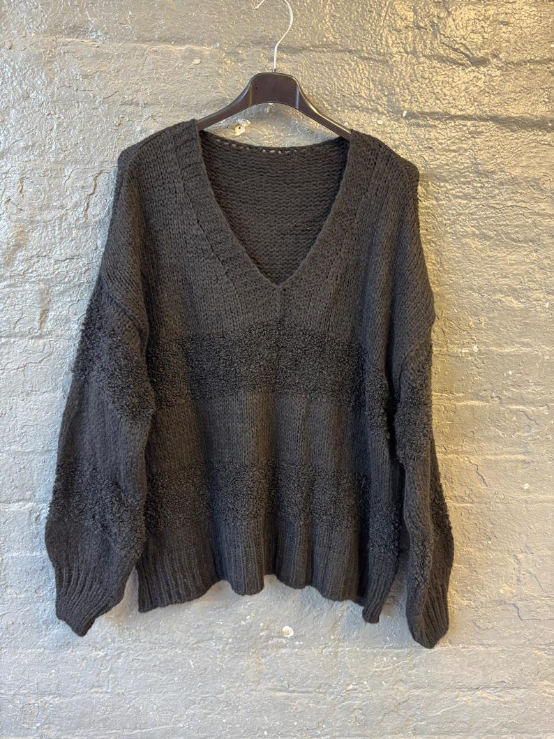 May include: A dark gray, oversized sweater with a deep V-neck. The sweater is hanging on a black hanger against a gray brick wall. The knitwear features a textured horizontal stripe design across the chest.