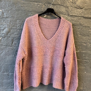May include: A mauve-colored, V-neck sweater with a textured knit design. The sweater is hanging on a black hanger against a dark gray brick wall. The sleeves and hem have a ribbed knit finish.