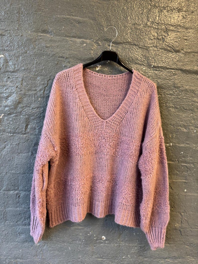 May include: A mauve-colored, V-neck sweater with a textured knit design. The sweater is hanging on a black hanger against a dark gray brick wall. The sleeves and hem have a ribbed knit finish.