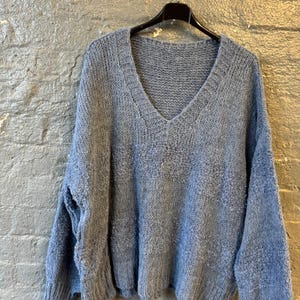 May include: A light blue, oversized sweater with a deep V-neck. The sweater is made of a soft, textured knit fabric and hangs on a black hanger against a gray brick wall. The sleeves are long and the fit is loose.