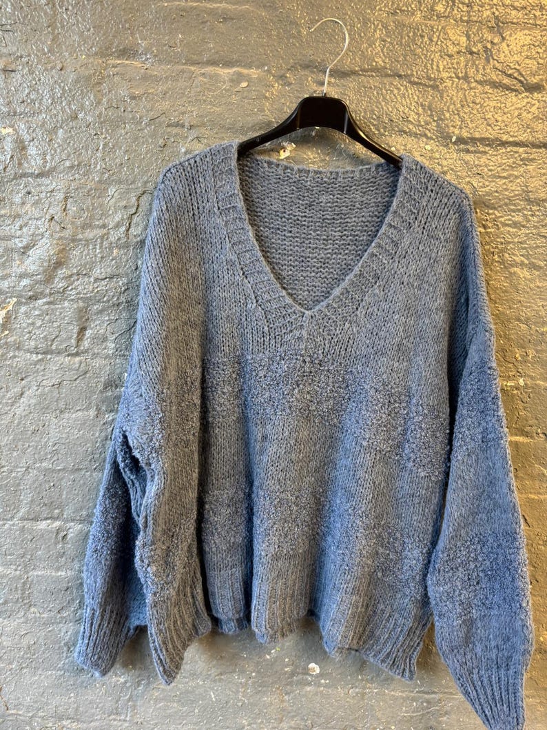 May include: A light blue, oversized sweater with a deep V-neck. The sweater is made of a soft, textured knit fabric and hangs on a black hanger against a gray brick wall. The sleeves are long and the fit is loose.
