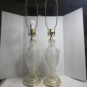 May include: A pair of clear glass table lamps featuring intricate pressed patterns, including diamond and floral motifs. Each lamp has a brass-toned metal socket, a tall wire harp for a shade, and a circular base with a white power cord.