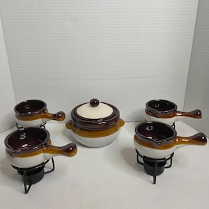 Könnte beinhalten: Ein Set aus Keramik-Suppenschalen und einer Servierschüssel mit Deckel. Die Schalen sind weiß mit braunem Rand und Griff und stehen auf schwarzen Metallständern. Die Servierschüssel ist braun und weiß mit passendem Deckel.