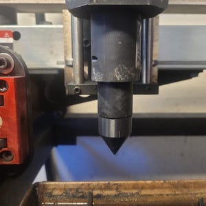 Op de afbeelding: Close-up van de spindel en de snijbit van een CNC-machine. De spindel is donkergrijs en cilindrisch, met een zwarte, conische snijbit die naar beneden uitsteekt. Rode en zilveren machineonderdelen zijn zichtbaar aan de linkerkant.