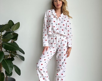 May include: A white long-sleeve pajama set featuring a button-down shirt with a collar and matching pants, both decorated with a repeating pattern of small red and grey hearts. The pants include an elastic waistband with a drawstring tie.
