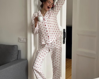 Op de afbeelding: Een witte pyjamaset met lange mouwen, bestaande uit een overhemd met knoopjes en een bijpassende lange broek, beide voorzien van een herhalend patroon van kleine rode hartjes. De set bevat een kraagshirt met rode knopen en een loszittende broek, gecombineerd met witte sokken.