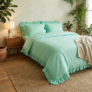 May include: A mint green bed set with ruffled edges, including a duvet cover and two pillowcases, is displayed on a bed. The bedroom features a jute rug, wooden nightstand, and potted plants, creating a serene and inviting atmosphere.