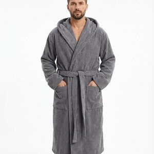 Könnte beinhalten: Ein grauer Bademantel mit Kapuze, passendem Gürtel und Taschen. Der Frotteebademantel ist knielang und hat eine lockere Passform. Das Model trägt den Bademantel.
