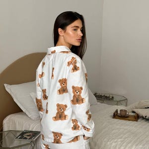 以下が含まれることがあります： 茶色のテディベアの繰り返し模様が特徴の白いパジャマセット。長袖トップスとパンツは柔らかい生地で作られています。パジャマは快適さとリラックスのために設計されています。