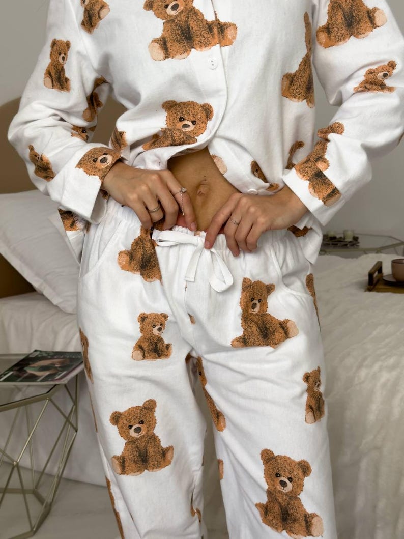 以下が含まれることがあります： 茶色のテディベアの繰り返し模様が特徴の白いパジャマセット。長袖トップスはボタンダウンフロントで、パンツはドローストリングウエストです。テディベアのデザインはセット全体に一貫しています。