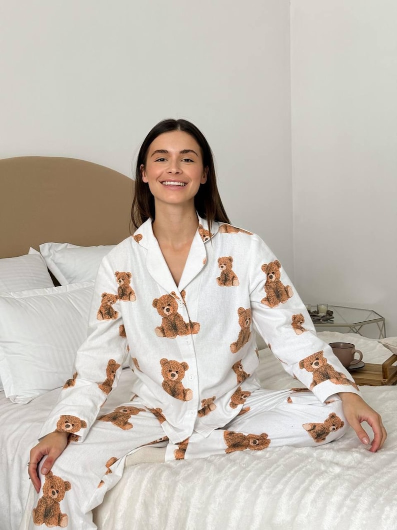 以下が含まれることがあります： 茶色のテディベアの繰り返し模様が特徴の白いパジャマセット。長袖トップスはボタンダウンフロントとノッチカラー。お揃いのパンツがスリープウェアアンサンブルを完成させ、居心地が良く遊び心のある外観を作り出しています。