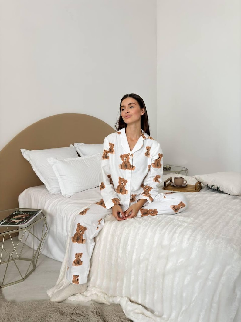 以下が含まれることがあります： 茶色のテディベアの繰り返し模様が施された白いパジャマセット。長袖トップスはボタンダウンフロントとノッチカラー。パンツは長くストレートレッグです。パジャマは柔らかく軽い生地で作られています。