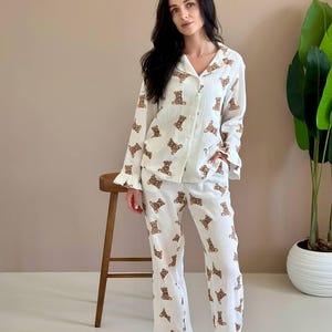 Könnte beinhalten: Ein weißer langärmeliger Schlafanzug mit einem sich wiederholenden braunen Teddybärenmuster. Das Oberteil hat einen Kragen mit Knöpfen, und sowohl das Oberteil als auch die lange Hose sind mit Rüschen an den Säumen versehen. Der Stoff ist strukturiert.