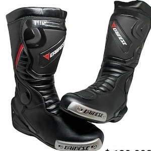 Puede incluir: Par de botas de moto negras con el nombre de la marca "DAINESE" en blanco. Las botas tienen detalles rojos y plateados. Las botas son de cuero y tienen un diseño protector.