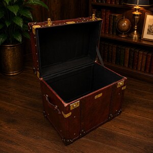May include: An open, antique-style trunk with a dark brown leather exterior and brass hardware. The interior is lined with black fabric. The trunk has a handle and is sitting on a wooden floor. The trunk is empty.