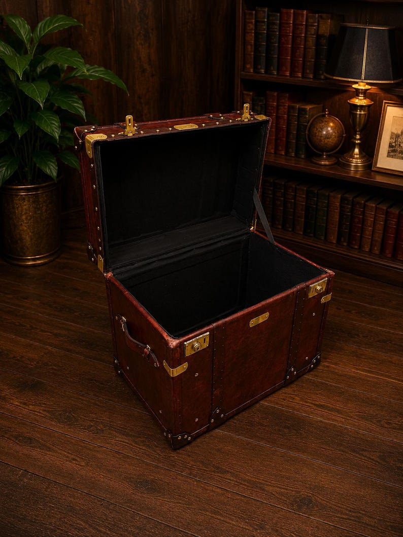 May include: An open, antique-style trunk with a dark brown leather exterior and brass hardware. The interior is lined with black fabric. The trunk has a handle and is sitting on a wooden floor. The trunk is empty.