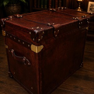 May include: A large, rectangular, dark brown leather trunk with brass corner protectors and rivets. The trunk has a leather handle and is set against a dark wood background. The trunk is a vintage style storage chest.