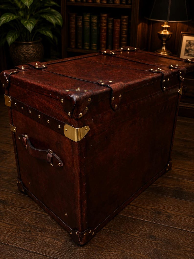 May include: A large, rectangular, dark brown leather trunk with brass corner protectors and rivets. The trunk has a leather handle and is set against a dark wood background. The trunk is a vintage style storage chest.