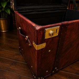 May include: A vintage-style, open trunk with a rich, reddish-brown leather exterior. The trunk features brass hardware, including corner protectors, a lock, and rivets. The interior is lined with black fabric, and a leather handle is visible on the side.