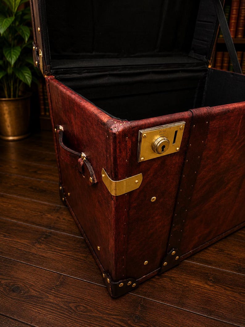 May include: A vintage-style, open trunk with a rich, reddish-brown leather exterior. The trunk features brass hardware, including corner protectors, a lock, and rivets. The interior is lined with black fabric, and a leather handle is visible on the side.