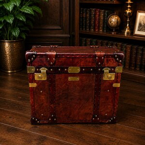 May include: A vintage-style, rectangular trunk made of dark brown leather. The trunk features brass hardware, including corner protectors, latches, and decorative studs. The trunk is a rich, reddish-brown color and appears to be well-crafted.