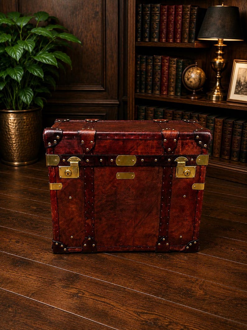 May include: A vintage-style, rectangular trunk made of dark brown leather. The trunk features brass hardware, including corner protectors, latches, and decorative studs. The trunk is a rich, reddish-brown color and appears to be well-crafted.