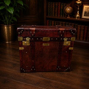 May include: A vintage-style, rectangular trunk made of dark brown leather with brass hardware. The trunk features metal studs, corner protectors, and brass locks. It is a classic piece of luggage or storage.