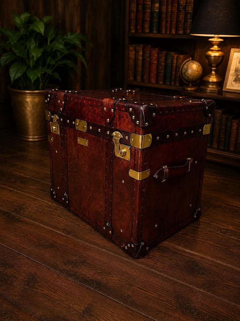 May include: A vintage-style, dark brown leather trunk with brass hardware, including corner protectors, clasps, and rivets. The trunk features a handle and is set on a wooden floor, suggesting a travel or storage theme.