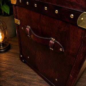 May include: A close-up of a vintage wooden trunk with a rich, dark brown finish. The trunk features brass accents, including corner protectors and decorative studs. A leather handle is attached to the front. The trunk is set on a wooden surface.
