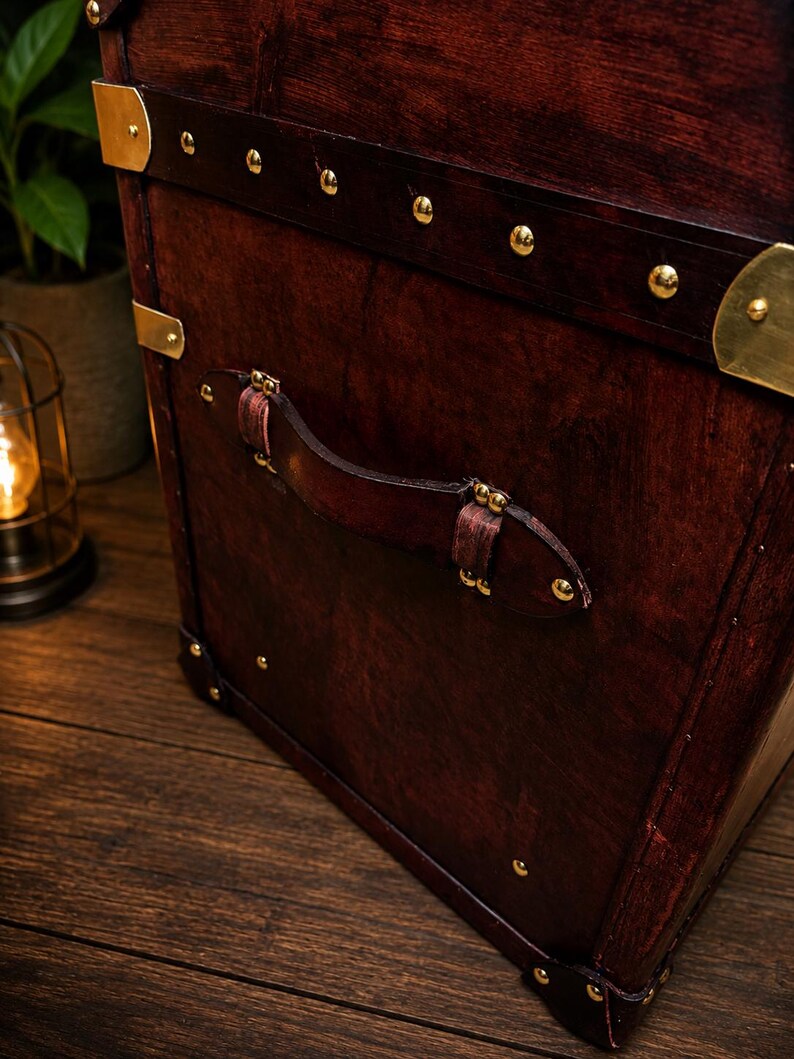 May include: A close-up of a vintage wooden trunk with a rich, dark brown finish. The trunk features brass accents, including corner protectors and decorative studs. A leather handle is attached to the front. The trunk is set on a wooden surface.