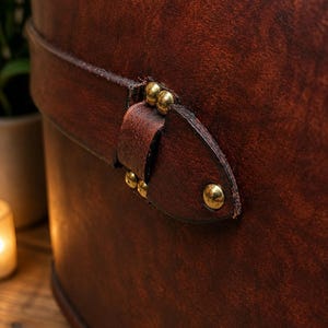 May include: Close-up of a brown leather container with a strap and brass rivets. The leather has a rich, textured appearance with variations in color. The strap is secured with small, round brass rivets.