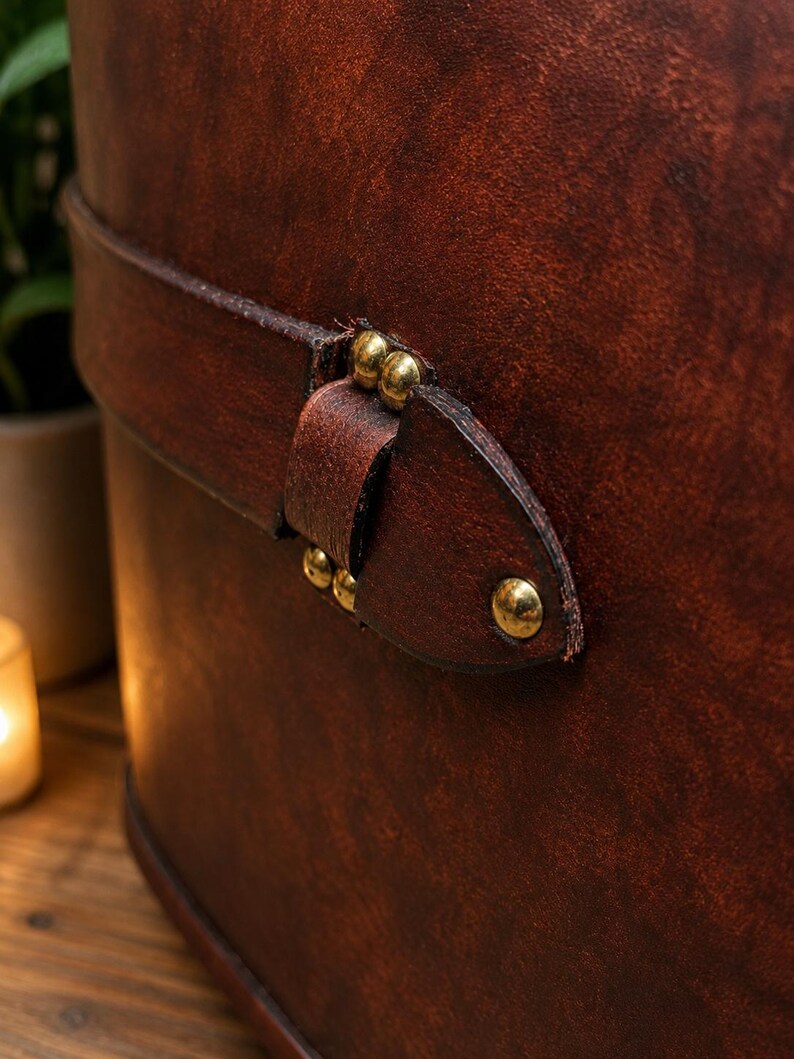 May include: Close-up of a brown leather container with a strap and brass rivets. The leather has a rich, textured appearance with variations in color. The strap is secured with small, round brass rivets.