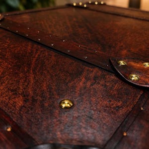 May include: Close-up of a dark brown wooden chest with brass-colored studs. The chest has a leather trim and a rich, textured wood grain. The details suggest a vintage or antique style, with a focus on craftsmanship.