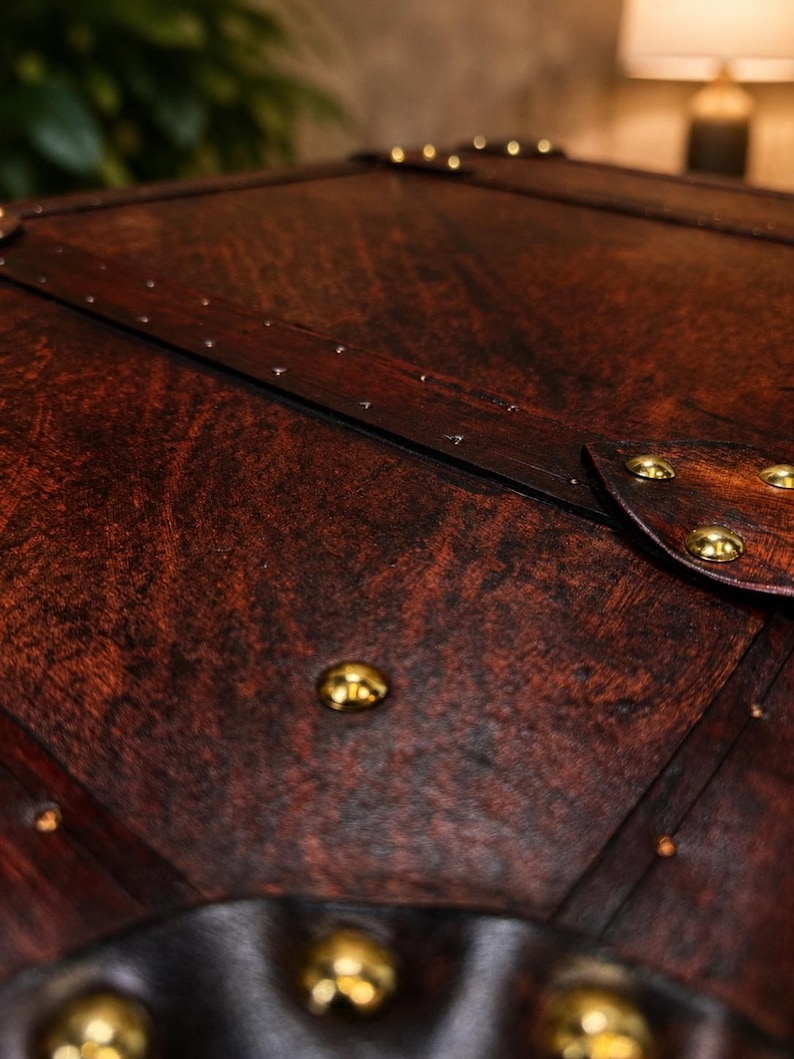 May include: Close-up of a dark brown wooden chest with brass-colored studs. The chest has a leather trim and a rich, textured wood grain. The details suggest a vintage or antique style, with a focus on craftsmanship.
