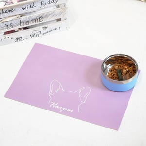 May include: A light purple pet placemat with a white outline of a dog's head and the name "Harper" in script. A stainless steel and light blue pet bowl filled with food sits on the right. A wooden crate with text is in the background.